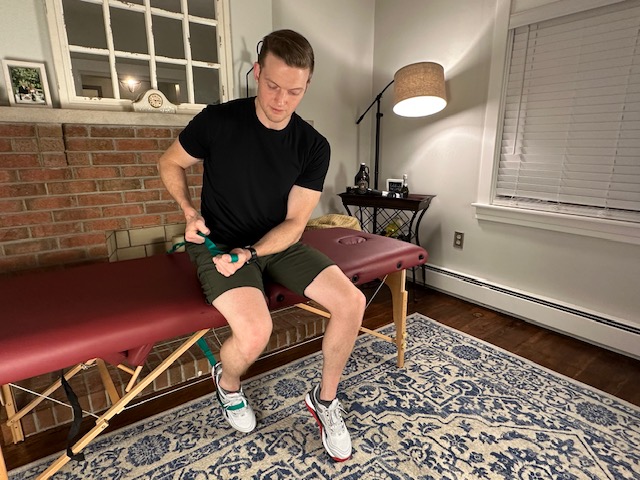 Demonstrating a seated knee bend with a strap on a massage table to improve knee flexion range of motion