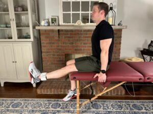 demonstrating long arc quad exercise with an ankle weight sitting on the edge of a massage table